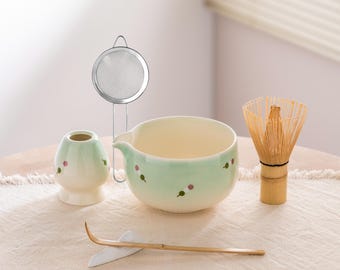 May include: A matcha tea set featuring a light green ceramic bowl and whisk, a small ceramic container, a bamboo scoop, and a stainless steel strainer. The bowl and container have a floral design.