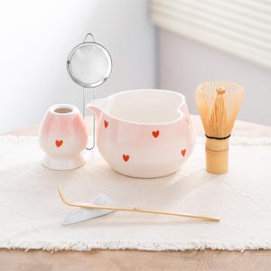 May include: A matcha tea set featuring a pink ceramic bowl with red hearts, a bamboo whisk, a bamboo scoop, a pink ceramic whisk holder, and a stainless steel strainer. The set is arranged on a white cloth.