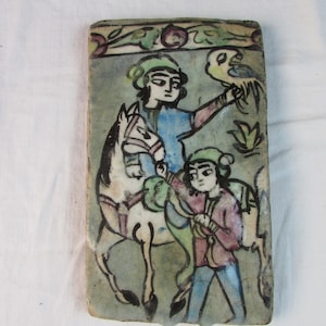 May include: A hand-painted ceramic tile depicting a scene with two figures. One figure is riding a white horse, while the other figure is standing and holding a bird. The tile has a green, blue, and white color scheme.