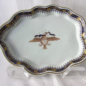May include: A white porcelain serving dish with a scalloped edge, featuring a brown and blue eagle design. The rim is decorated with gold and blue floral patterns. The dish is approximately 10 inches long.