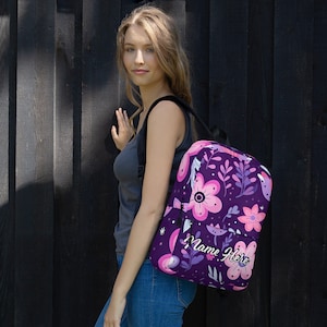 Puede incluir: Mochila morada con un estampado floral en rosa y blanco. La mochila tiene un bolsillo con cremallera y el texto "Name Here" está impreso en la parte delantera.