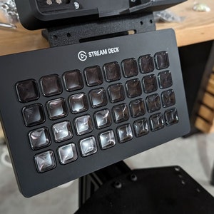 May include: A black Stream Deck with 32 customisable buttons. The buttons are clear and have a slight shine. The Stream Deck is a popular tool for streamers and content creators.