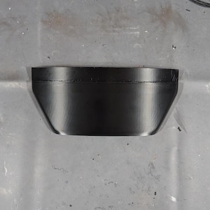 May include: A black, trapezoidal-shaped object with a smooth, reflective surface. The object has a slightly curved top edge and angled sides, giving it a modern, minimalist appearance. The object appears to be made of a solid material.