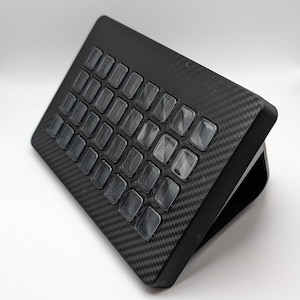 May include: A black keyboard with a carbon fiber pattern and clear plastic keys. The keyboard is angled up and has a stand on the back.