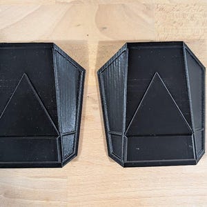 May include: Two black, geometrically shaped, 3D printed objects. Each object has a triangular shape in the center and a slightly rounded, octagonal outer edge.