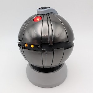 May include: A dark grey, spherical, decorative object with a textured surface and a small grey handle on top. The object has three small, orange lights and one red light on the front. The object is sitting on a grey base.