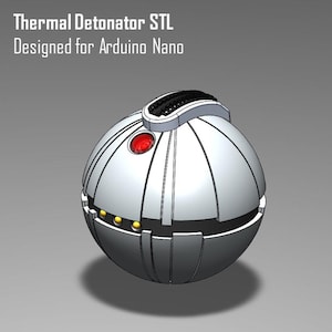 Pode incluir: Um dispositivo esférico prateado com um botão central vermelho e uma secção superior preta. O texto "Thermal Detonator STL Designed for Arduino Nano" é exibido no topo. O dispositivo tem uma série de linhas verticais e pequenos detalhes dourados.