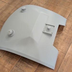 Può includere: Un coperchio in plastica grigia con tre pomelli rotondi e un ritaglio rettangolare. Il coperchio ha una forma curva e una superficie liscia.