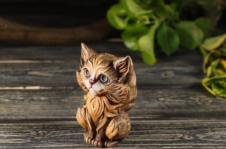 Pu&ograve; includere: Una statuetta di legno di un gatto marrone e bianco con grandi occhi. Il gatto &egrave; seduto con la testa inclinata di lato e la coda arrotolata attorno al suo corpo.