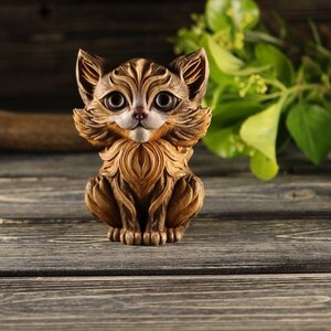 Pu&ograve; includere: Una statuetta di legno di un gatto con pelliccia marrone e grandi occhi rotondi. Il gatto &egrave; seduto con le zampe ripiegate sotto il corpo e ha un'espressione amichevole.