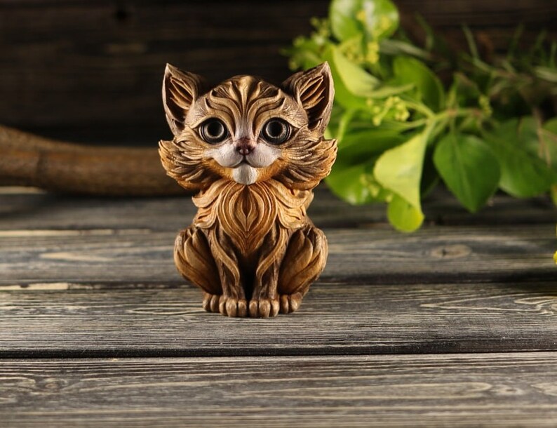 Pu&ograve; includere: Una statuetta di legno di un gatto con pelliccia marrone e grandi occhi rotondi. Il gatto &egrave; seduto con le zampe ripiegate sotto il corpo e ha un'espressione amichevole.