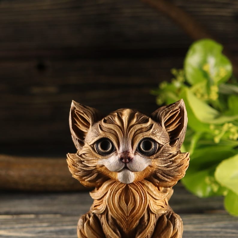 Pu&ograve; includere: Una statuetta di legno di un gatto con pelliccia marrone e grandi occhi espressivi. Il gatto &egrave; seduto con le zampe ripiegate sotto di lui e ha un'espressione amichevole.