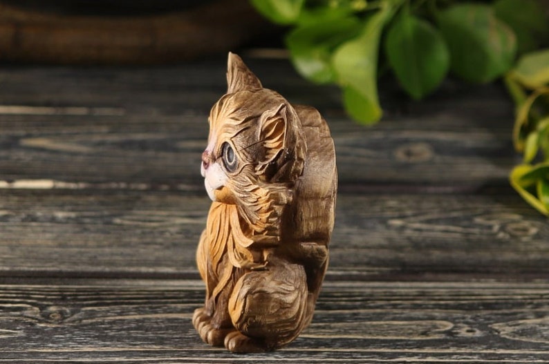 Pu&ograve; includere: Una statuetta di legno intagliata a mano di un gatto marrone e bianco seduto con la testa girata di lato. Il gatto ha un viso dettagliato con occhi dipinti e una coda folta.