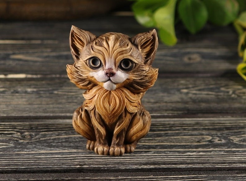 Pu&ograve; includere: Una piccola statuetta di legno intagliata in modo intricato che rappresenta un gatto marrone e bianco con grandi occhi espressivi. Il gatto &egrave; seduto con le zampe ripiegate sotto di lui e la coda arrotolata attorno al suo corpo.