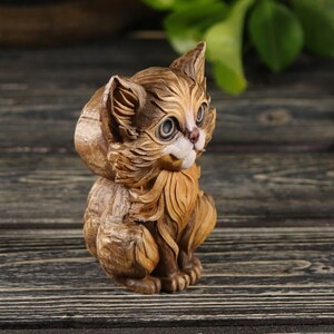 Pu&ograve; includere: Una statuetta di legno intagliata a mano di un gatto seduto con la testa inclinata di lato. Il gatto &egrave; marrone e ha il viso bianco con gli occhi neri.
