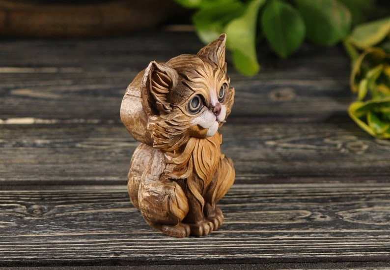 Pu&ograve; includere: Una statuetta di legno intagliata a mano di un gatto seduto con la testa inclinata di lato. Il gatto &egrave; marrone e ha il viso bianco con gli occhi neri.