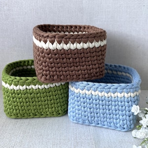 May include: Three handmade crochet baskets in brown, green, and blue. Each basket has a white stripe around the top. The square-shaped baskets are made of thick yarn, suitable for storage or decoration. The measurements are in cm.