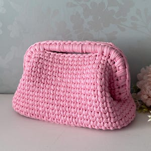 May include: A light pink crochet handbag featuring a structured frame opening and a thick, textured knit pattern. The bag has a rectangular base and rounded sides, constructed from a continuous cord-like yarn in a solid pastel pink colour.