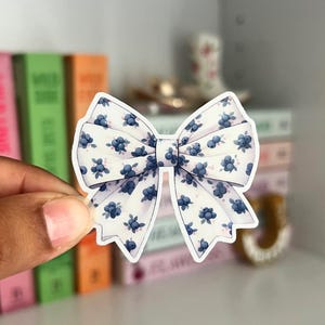 May include: A white bow-shaped sticker with a blue floral pattern. The sticker is held in front of a shelf with books and decorative items. The bow has a delicate, feminine design.
