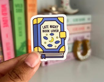 May include: A sticker featuring a blue book design with the text "LATE NIGHT BOOK LOVER". The sticker has a yellow border and includes illustrations of a moon, stars, and clouds. The sticker is held in front of a shelf with books.