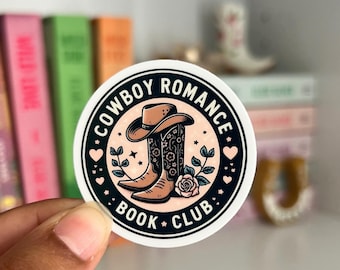 Op de afbeelding: Een ronde sticker met de tekst "COWBOY ROMANCE BOOK CLUB" rondom een ontwerp van een cowboyhoed en -laarzen. De laarzen zijn versierd met bloemmotieven. De sticker wordt in een hand gehouden, met boeken op de achtergrond.