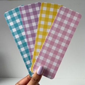 May include: Four bookmarks with a gingham pattern in teal, purple, yellow, and pink. Each bookmark has a white grid pattern. The bookmarks are held in a hand with light purple nail polish.