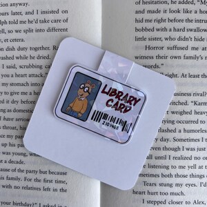 Puede incluir: Un marcap&aacute;ginas colorido con un personaje de dibujos animados del programa infantil "Arthur." El marcap&aacute;ginas tiene un acabado hologr&aacute;fico y dice "Library Card" con un c&oacute;digo de barras que dice "2101408."