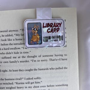 Puede incluir: Un personaje de dibujos animados con gafas y una camisa marr&oacute;n sostiene una tarjeta de biblioteca con el texto "LIBRARY CARD" y el n&uacute;mero "2101908".