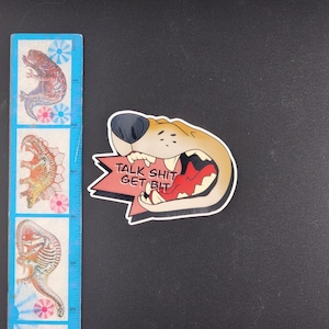 May include: A sticker featuring a cartoon dog's open mouth with the text "TALK SHIT GET BIT" on a red and white starburst. A blue ruler with dinosaur illustrations is on the left.