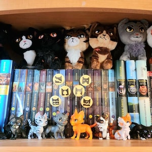 Puede incluir: Una colecci&oacute;n de art&iacute;culos de la serie de libros Warriors que incluye peluches de gatos en el estante superior y peque&ntilde;as figuras de gatos en el inferior. Los estantes est&aacute;n llenos de lomos de libros con el t&iacute;tulo Warriors e ilustraciones.