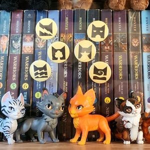 Puede incluir: Cuatro figuras de gatos en colores blanco, gris, naranja y carey se sit&uacute;an frente a una fila de lomos de libros Warriors. Sobre las figuras, seis pines circulares amarillos muestran s&iacute;mbolos negros de un rayo, un &aacute;rbol y olas.