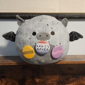 May include: A round, gray, speckled plush toy shaped like a bat with small ears and black wings. It features three circular pinback buttons attached to the front: a yellow one reading NERVOUS, a white one reading RABIES HAVER, and a purple one reading FurFag.