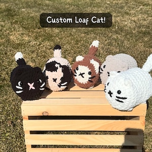 May include: Five handmade crochet cat toys in various colors, including black, white, brown, and beige, are displayed on a wooden crate. Each cat has a unique face design and a small tail. The text "Custom Loaf Cat!" is above the cats.