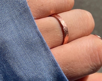 Puede incluir: Un anillo metálico fino con una textura martillada distintiva y un tono cobrizo rojizo cálido. La banda presenta una superficie reflectante que capta la luz, mostrando las hendiduras irregulares hechas a mano en toda su circunferencia.