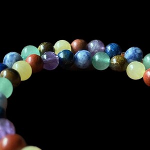 Könnte beinhalten: Nahaufnahme eines Armbands mit einer Vielzahl farbiger Edelsteine. Die runden, glatten Perlen sind in den Farbtönen Blau, Grün, Gelb, Lila und Braun gehalten. Das Armband ist vor einem schwarzen Hintergrund platziert.