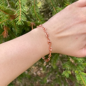 May include: A copper bracelet with a delicate, wavy link design. The bracelet is worn on a wrist, showcasing its intricate pattern and warm metallic hue. The background features green foliage.