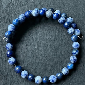 May include: A beaded bracelet featuring round, blue sodalite stones. The bracelet is strung on a silver-colored wire and has a circular shape. The stones vary in shades of blue and white, creating a marbled effect. The bracelet is displayed on a dark surface.