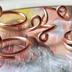 Puede incluir: Varios anillos de color cobre con un diseño único y arremolinado. Los anillos están hechos de un alambre grueso que ha sido doblado en bucles y curvas. Se muestran sobre un fondo colorido y abstracto. Los anillos son probablemente hechos a mano.