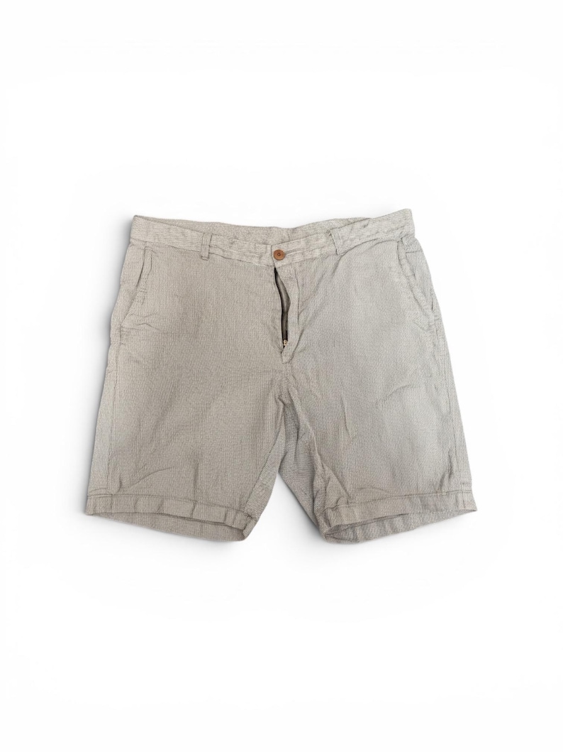 May include: Light beige shorts with a textured pattern. The shorts feature a button and zip closure, and front pockets. The garment is made of a lightweight fabric, suitable for warm weather.