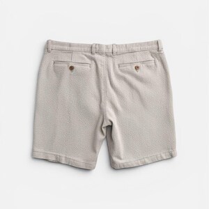 May include: Light beige shorts with a textured pattern. The shorts have two back pockets with brown buttons and a belt loop waistband. The shorts are made of a soft, woven fabric.