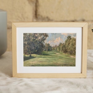 May include: A framed painting of a golf course with green grass, trees, and a blue sky with white clouds. The frame is made of light wood.