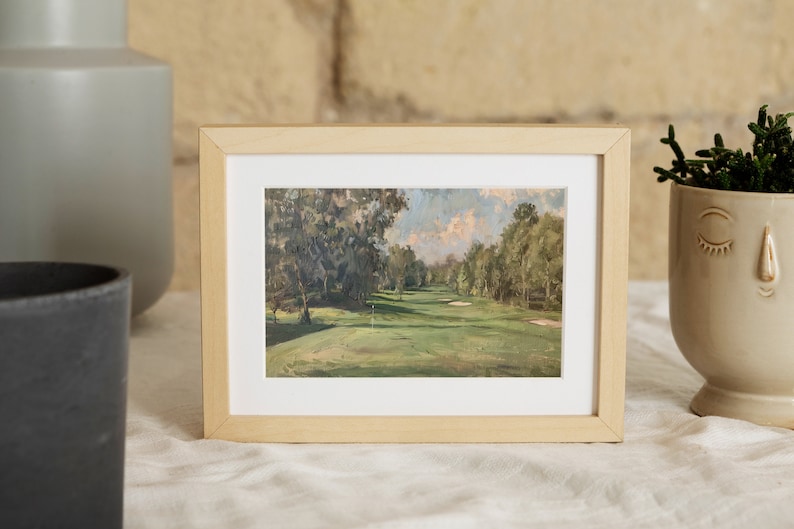 May include: A framed painting of a golf course with green grass, trees, and a blue sky with white clouds. The frame is made of light wood.
