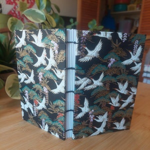 May include: A black and gold journal with a white crane pattern. The journal is closed and standing upright on a wooden surface.