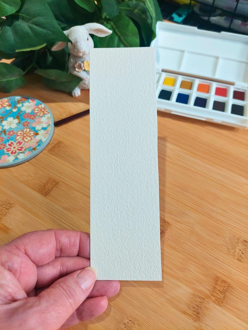 May include: A white watercolor paper bookmark with a textured surface.