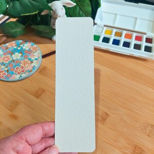 May include: A white bookmark with a textured surface. The bookmark is being held in a hand.