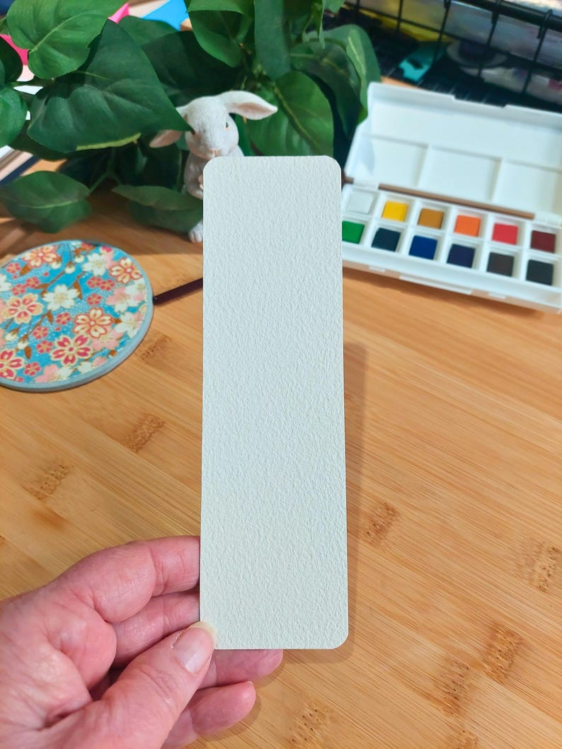 May include: A white bookmark with a textured surface. The bookmark is being held in a hand.