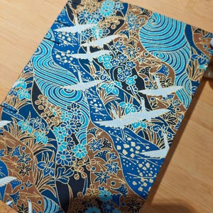 May include: A rectangular fabric with a Japanese-inspired design. The pattern features white cranes in flight, blue water waves, and floral elements in shades of blue, brown, and gold. The fabric is set against a wooden surface.