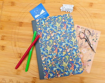 May include: A blue journal with a floral pattern, a red pen, a green pen, and a pair of decorative scissors are arranged on a wooden surface. A small blue card and a sticker that says "Take Time to Make Happy" are also present.