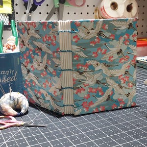 May include: Handmade journal with a cover featuring a crane and floral pattern in shades of blue, white, and pink. The journal is bound with visible stitching along the spine. The journal is sitting on a black surface.