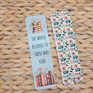 May include: Two rectangular bookmarks. One features a stack of books with flowers and the text "THE WORLD BELONGS TO THOSE WHO READ." The other has a floral pattern with pink flowers and green leaves. The background is a woven texture.